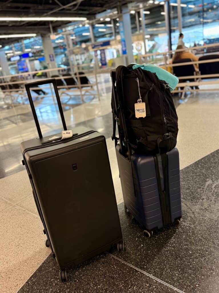 Two rolling suitcases, one black and one navy, stand upright in an airport terminal. A black backpack and a turquoise item rest on the navy suitcase, ready for a trip—perfect for anyone seeking a reliable Level8 review.