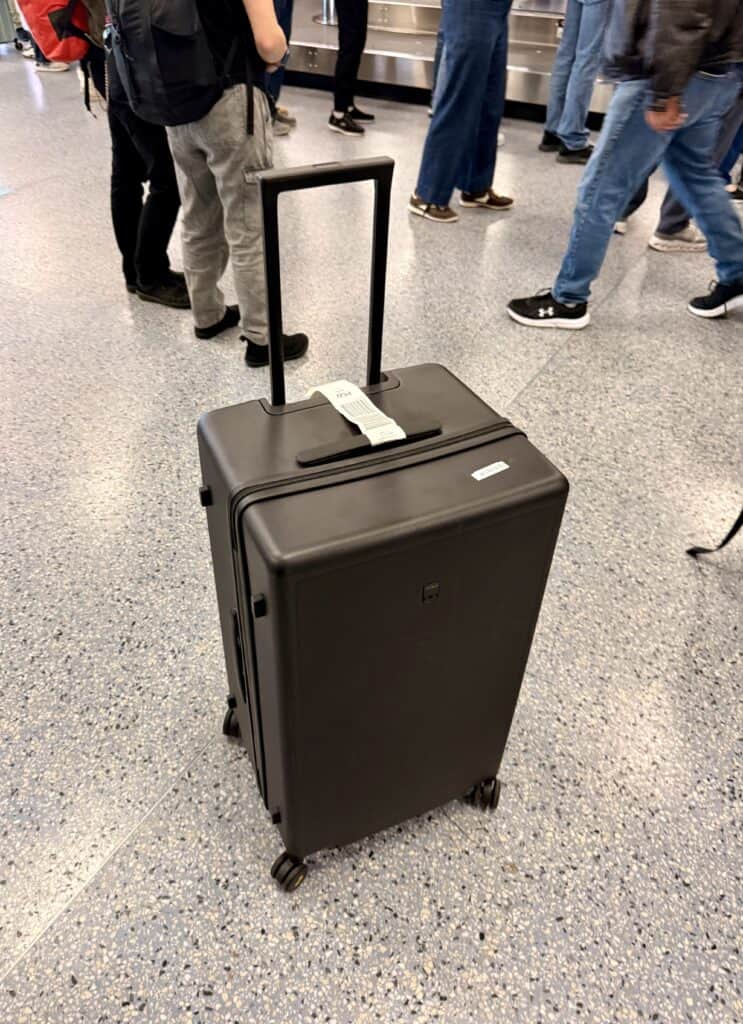 A black hard-shell suitcase with an airline baggage tag stands upright on a speckled airport floor near people waiting, making it a prime candidate for any level8 luggage review.
