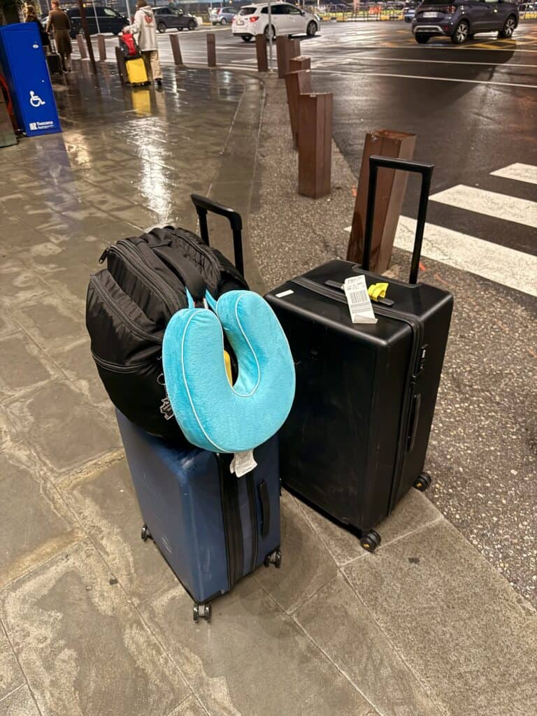 Two suitcases and a black backpack with a blue travel neck pillow sit on a wet sidewalk near a street and crosswalk at night, perfect for anyone considering writing a level8 luggage review.