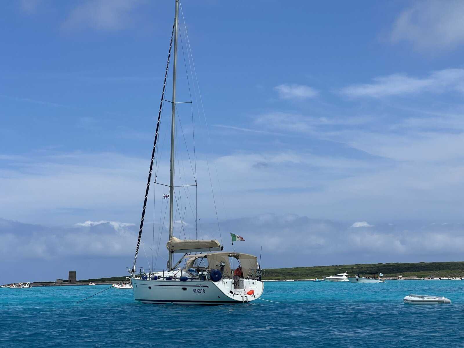 A white sailboat with an Italian flag floats on turquoise water near a low, green shoreline under a partly cloudy sky.
