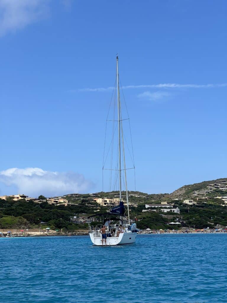 A white sailboat is anchored in calm blue water near a coastline with green hills and scattered houses under a clear blue sky, capturing the tranquil charm of Alghero Sardinia—one of the top things to do in Alghero.