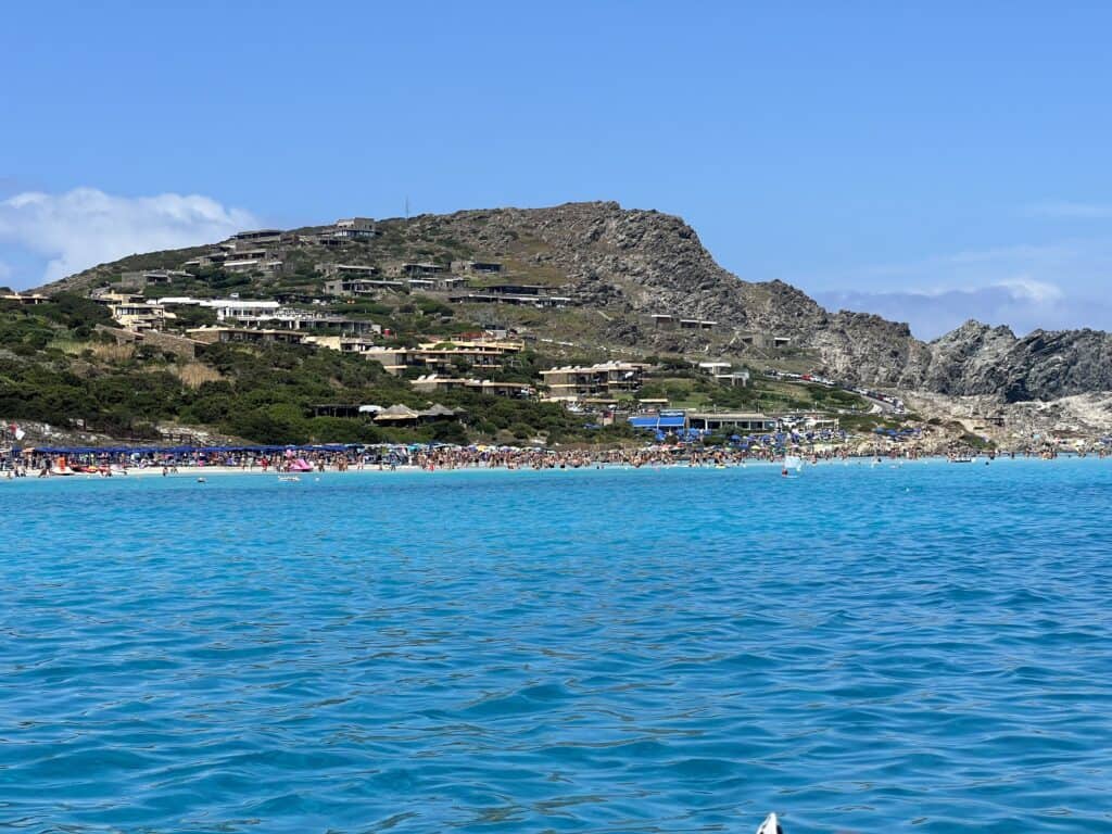 Bright blue sea with clear water in the foreground, a sandy beach crowded with people, and rocky hills with houses in the background under a clear sky—perfect for discovering things to do in Alghero Sardinia.