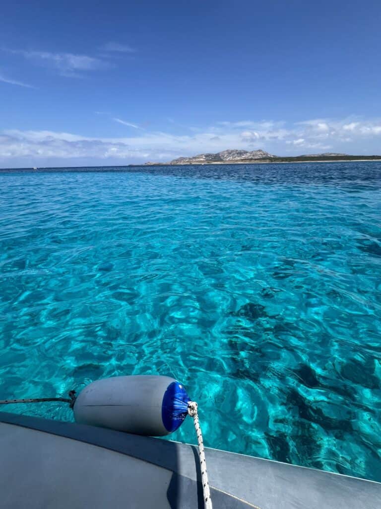 Clear turquoise water view from a boat with a blue and white fender in the foreground; coastline and distant hills under a blue sky—an inviting scene highlighting why exploring by boat is among the top things to do in Alghero, Sardinia.