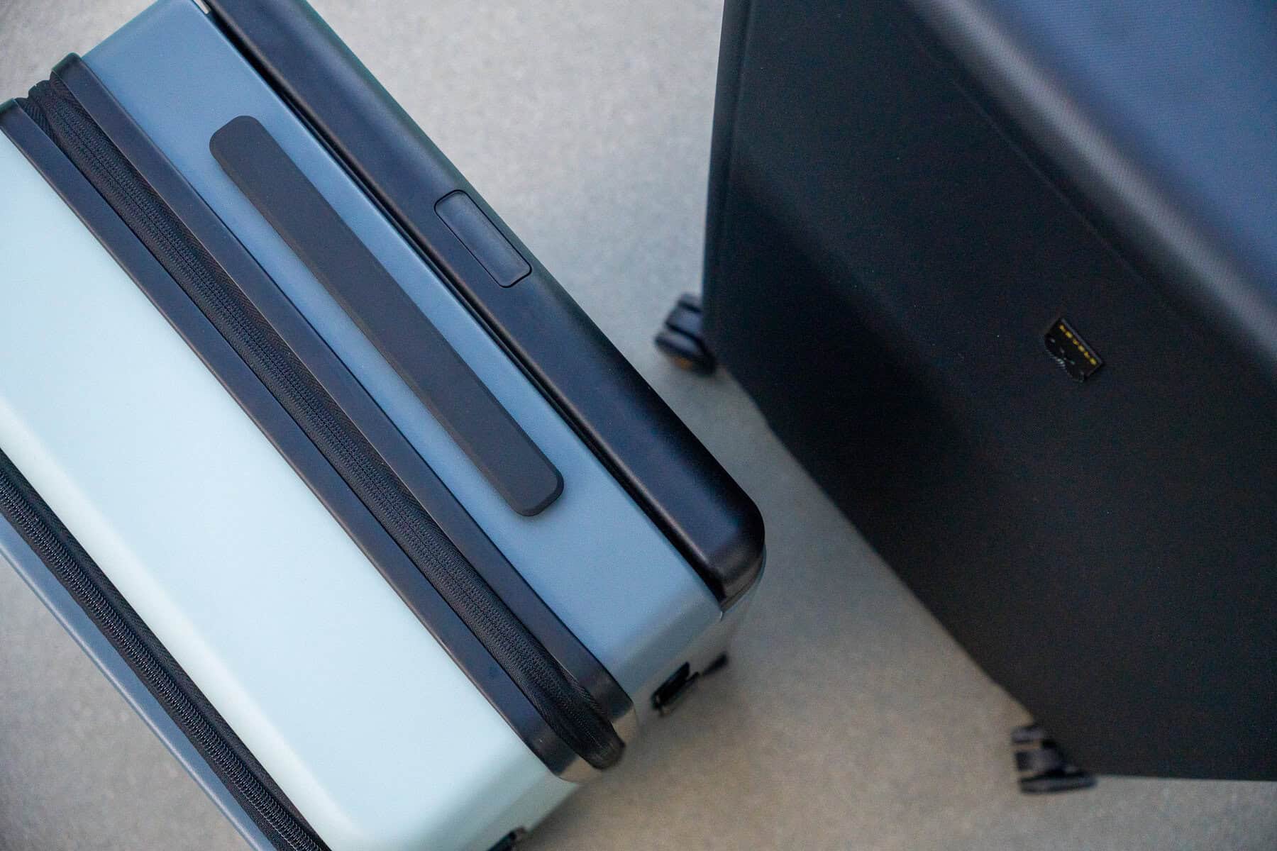 Two hard-shell suitcases, one light blue and one black, are pictured upright on a light gray surface, viewed from above.