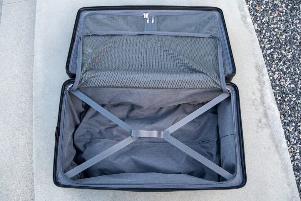 An empty, open suitcase with gray interior straps and a mesh pocket sits on a concrete surface next to gravel—perfect for those considering a Level8 luggage review.