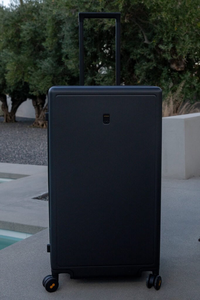 A large black hard-shell suitcase with an extended handle and four wheels is positioned upright outdoors near a concrete surface and greenery, perfect for anyone seeking real-life details for their Level8 luggage review.