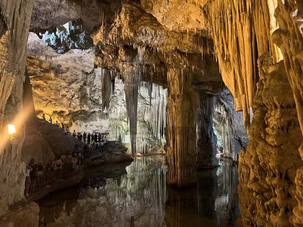 A large cave with stalactites and stalagmites, reflected in a pool of water, features people walking along a lit pathway inside—one of the top things to do in Alghero, Sardinia for nature and adventure lovers.