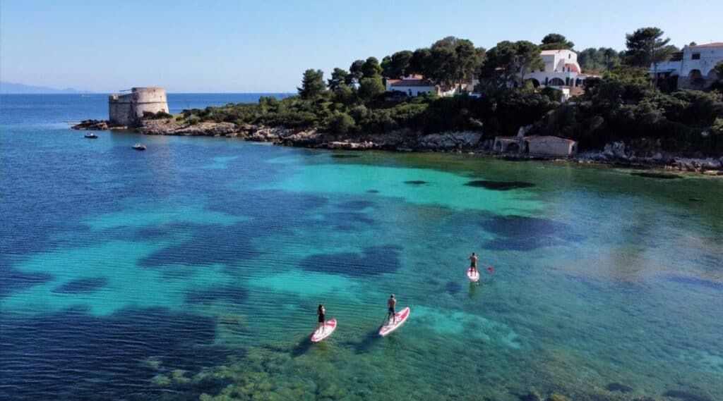 Three people paddleboard on clear turquoise water near a rocky shoreline with old white buildings and trees in the background—one of the top things to do in Alghero, Sardinia.