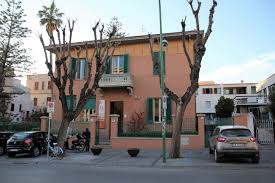 Two-story orange house with green shutters and a small balcony, bordered by pruned trees and a low fence, near popular things to do in Alghero Sardinia, with cars parked along the street in front.