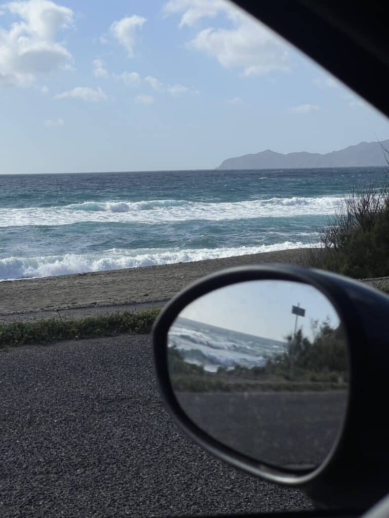 A view of a beach and ocean waves along the distant coastline of Alghero Sardinia, seen through a car window and reflected in the side mirror.