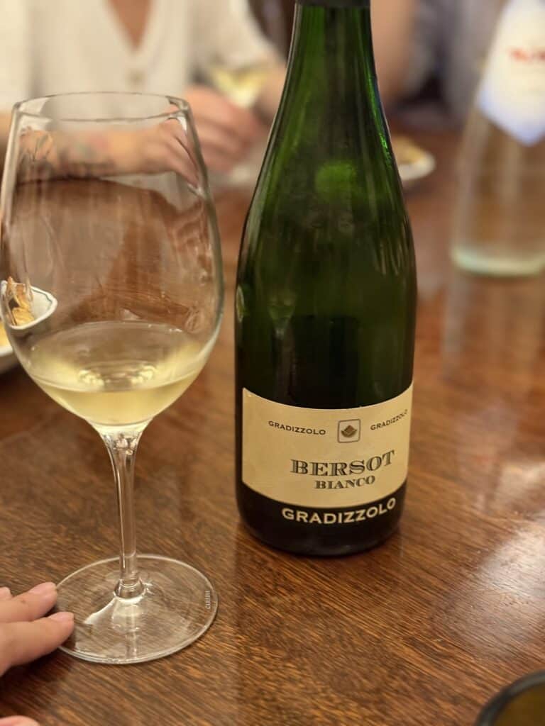 A bottle of Bersot Bianco Gradizzolo white wine and a filled glass sit on a wooden table, capturing the essence of a Bologna food tour, with blurred people in the background.