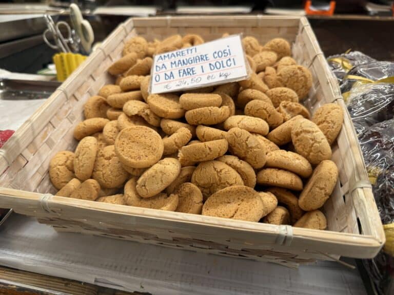 A basket filled with round amaretti cookies is displayed for sale with a handwritten sign in Italian indicating price per kilogram.