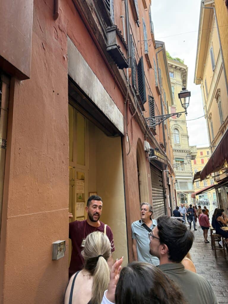 A small group of people stands in line outside a narrow entrance on a busy street lined with old buildings, ready to embark on a Bologna food tour.