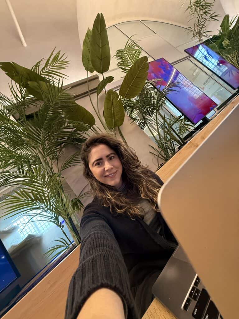 Woman sitting at a desk with a laptop, taking a selfie in a modern office space with large green plants in the background, reviewing her language learning plan.