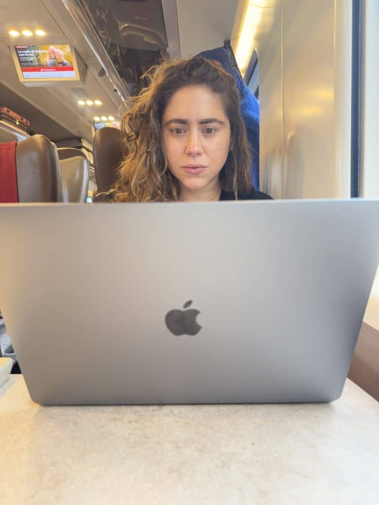 A person with curly hair works on a MacBook laptop while sitting at a table on a train, crafting a language learning plan, with empty seats and a screen visible in the background.