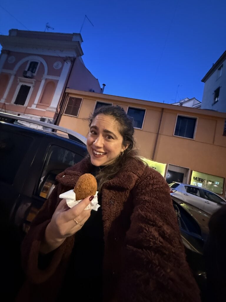 A woman wearing a brown coat holds a fried snack and smiles at the camera while standing on a street with colorful buildings at dusk, perhaps pausing during her travels to reflect on her language learning plan.