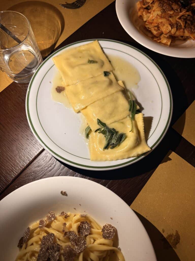 Three plates of pasta dishes on a table, perfect for discussing your next language learning plan—ravioli with sauce and greens, tagliatelle with truffle slices, and pasta with meat sauce. A glass of water sits close by.