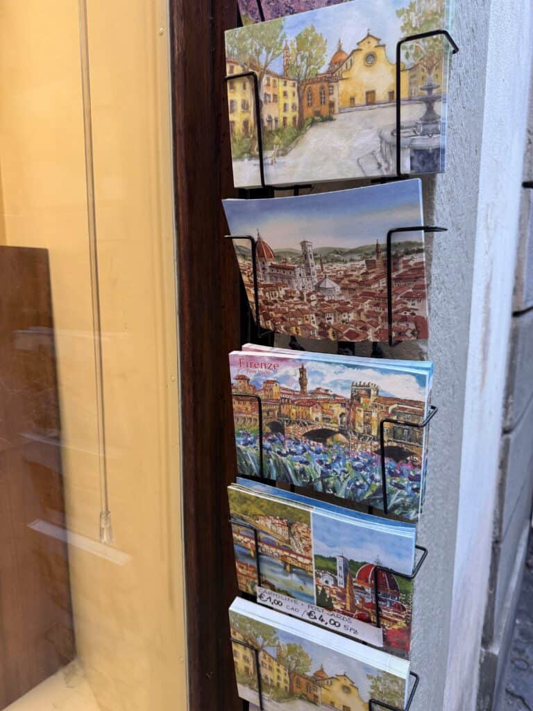 A display rack holds four rows of illustrated postcards featuring scenic city views and landmarks—perfect inspiration for your next language learning plan, positioned outside on a lively city street.