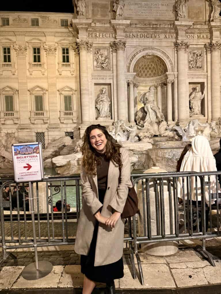 A woman in a tan coat stands smiling in front of the Trevi Fountain in Rome at night, holding her language learning plan, with a ticket sign and the fountain’s statues visible behind her.