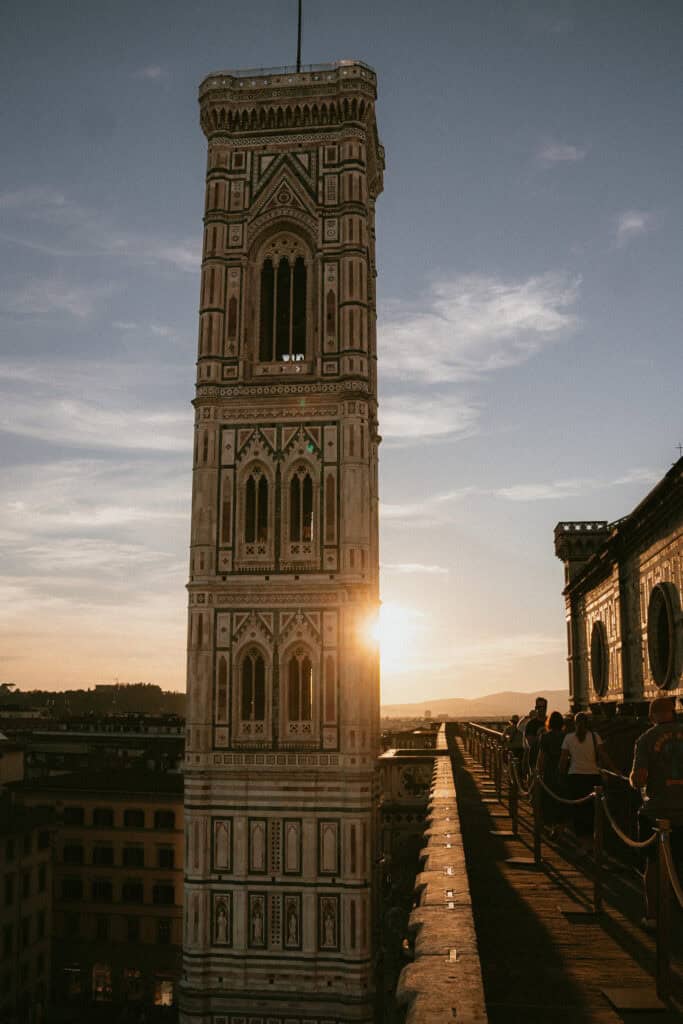 The photo shows Giotto’s Campanile, a tall, ornate bell tower in Florence, Italy, with the sun setting behind it and people walking along a nearby pathway after a day of truffle hunting in Florence.