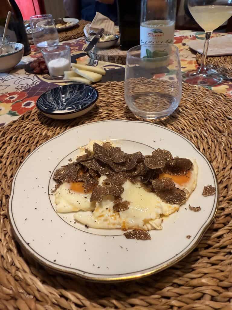A plate of fried eggs topped with sliced truffles, reminiscent of truffle hunting in Florence, sits on a woven placemat amid glasses, a wine bottle, and other dishes on a colorful tablecloth.