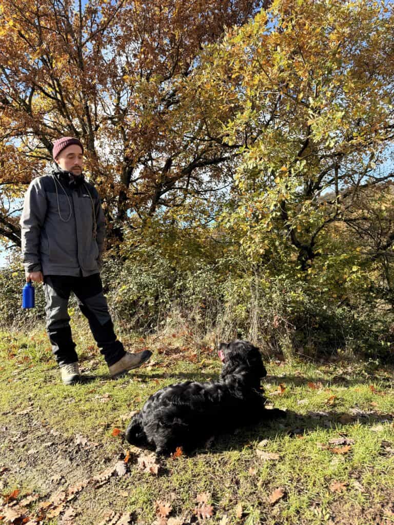 A person wearing outdoor clothing stands on a grassy area near autumn trees, perhaps during truffle hunting in Tuscany, looking at a black dog lying on the ground.