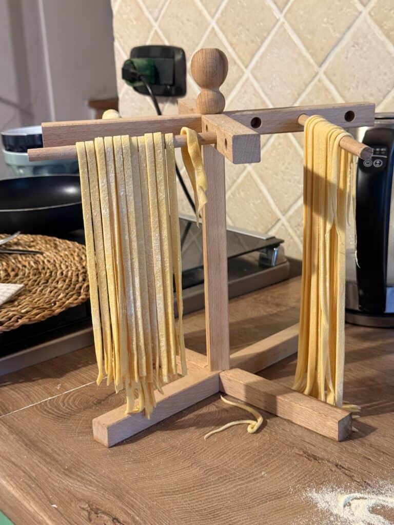Fresh pasta noodles hang on a wooden drying rack in a cozy kitchen, reminiscent of afternoons spent truffle hunting in Tuscany, with appliances and a woven placemat adding to the authentic Italian charm.