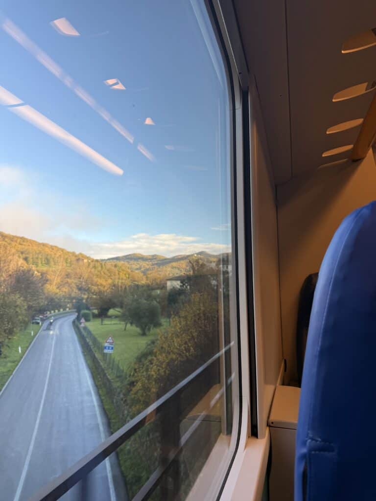 View from a train window showing a winding road, green hills perfect for truffle hunting in Tuscany, and distant mountains under a clear sky. The blue seat and part of the train interior are visible.