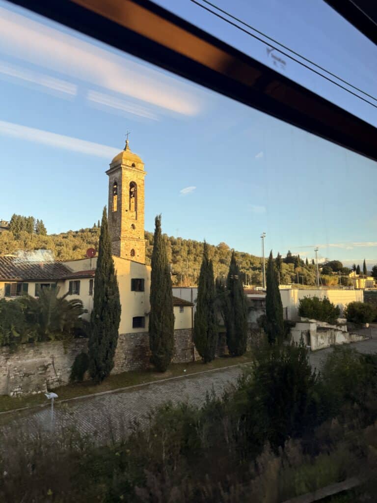 View from a train window showing a church bell tower, tall cypress trees, and tiled-roof buildings against hills—just the sort of scenery you might spot before truffle hunting in Tuscany under a clear sky.