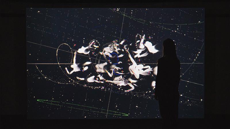 A silhouette of a person stands inside Casa Batlló, in front of a large digital display showing abstract white figures superimposed on a starry, celestial map.