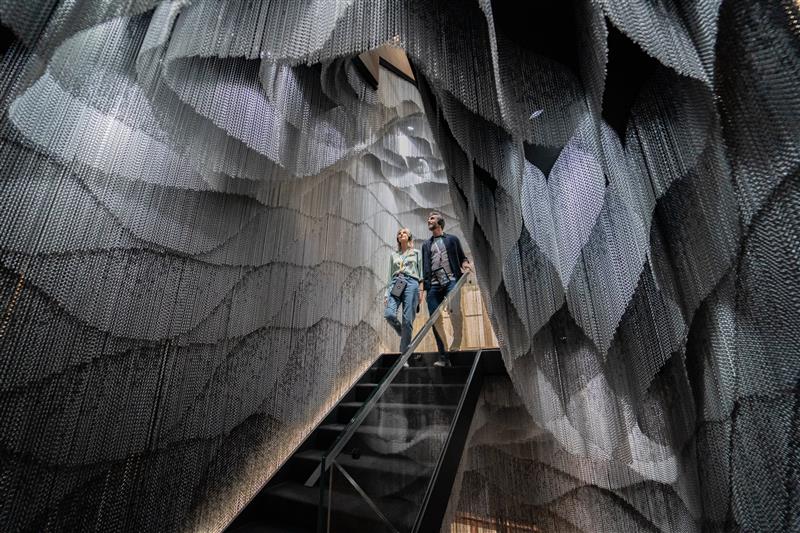 Two people descend a staircase inside Casa Batlló, surrounded by metallic, draped chain curtains that create a layered, cave-like effect.