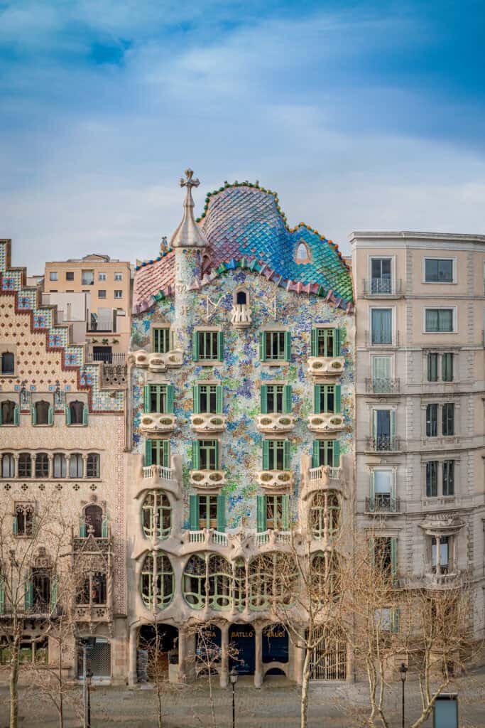 Ornate building with colorful, wavy facade and mosaic tiles, Casa Batlló stands out as it is flanked by more traditional structures under a blue sky.