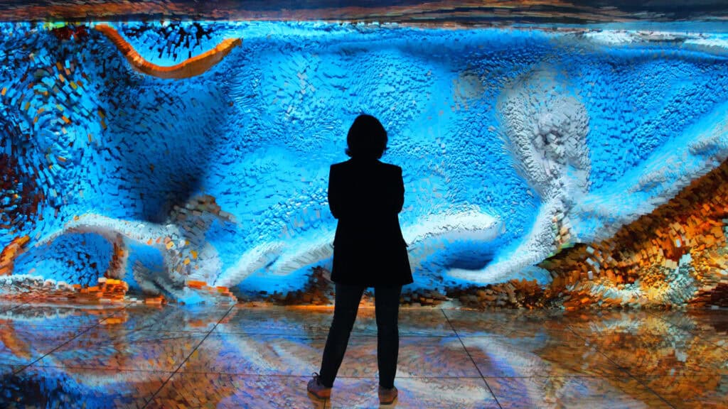 A person stands inside Casa Batlló, in front of a large, vibrant blue digital art display with abstract shapes and patterns, reflected on the shiny floor.