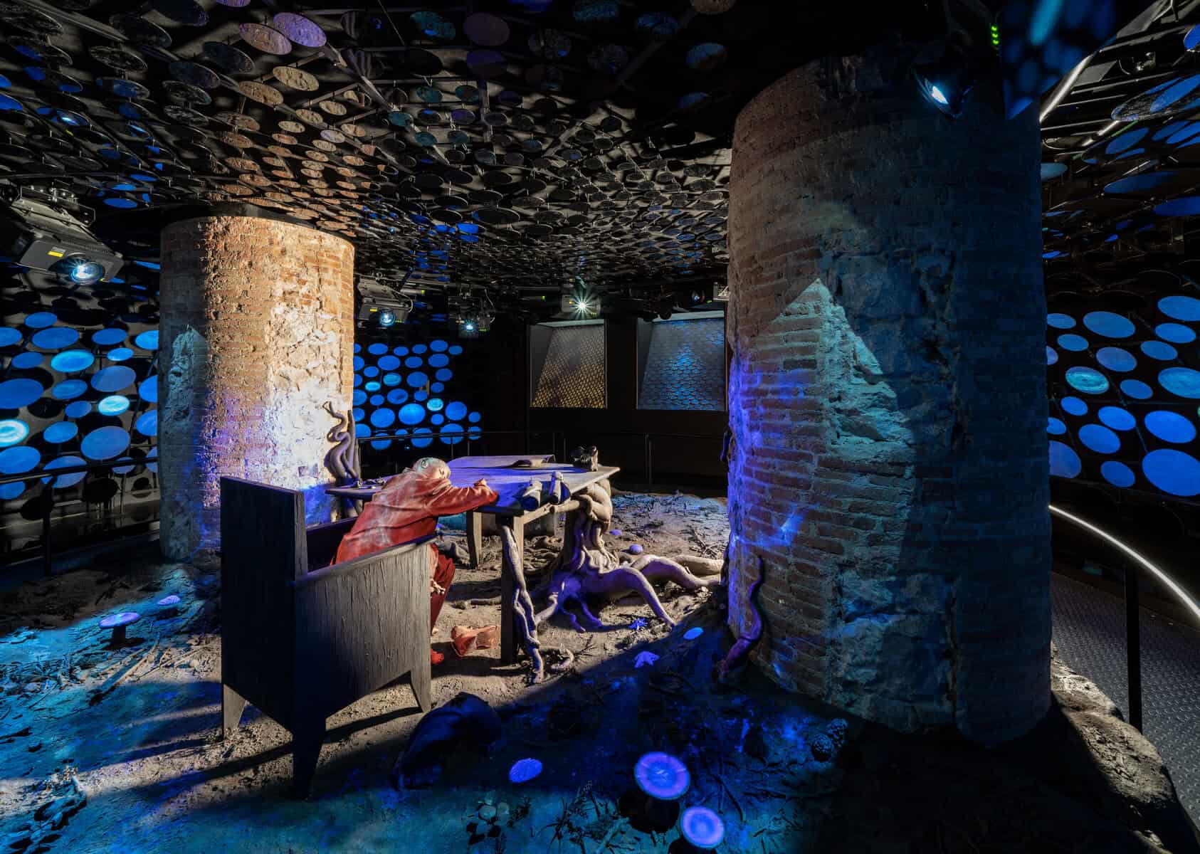 A dimly lit room inside Casa Batlló with brick columns, a seated human figure at a desk, tree roots winding across the floor, and blue circular lights softly illuminating the walls and ceiling.