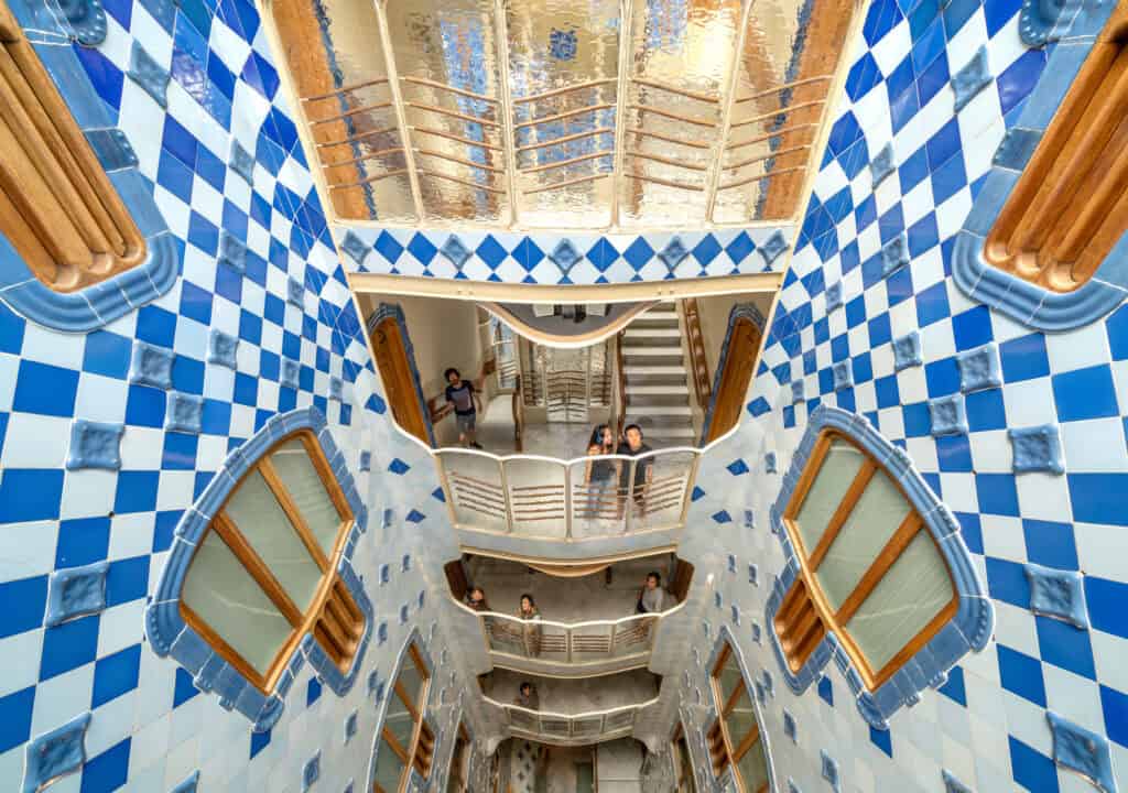 Inside Casa Batlló, the interior courtyard of the building features blue and white tile walls, wooden windows, and people looking over decorative balconies.
