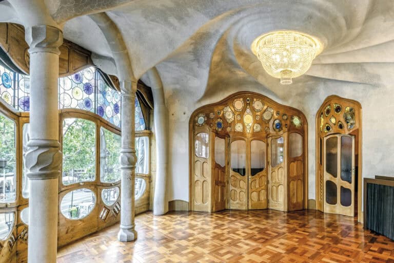 Ornate room with stained glass windows, curved wooden doors, a chandelier, and patterned parquet flooring in a modernist architectural style, reminiscent of the stunning interiors found inside Casa Batlló.