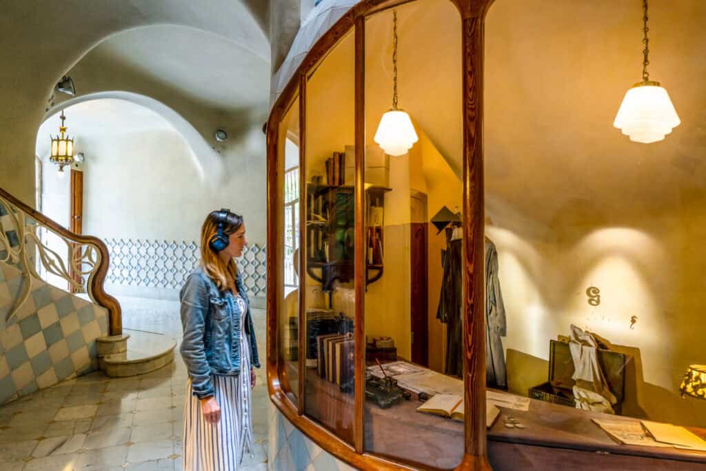 A woman wearing headphones stands in front of a vintage reception desk with papers and office items, inside Casa Batlló—a historic building with curved architecture and tiled walls.