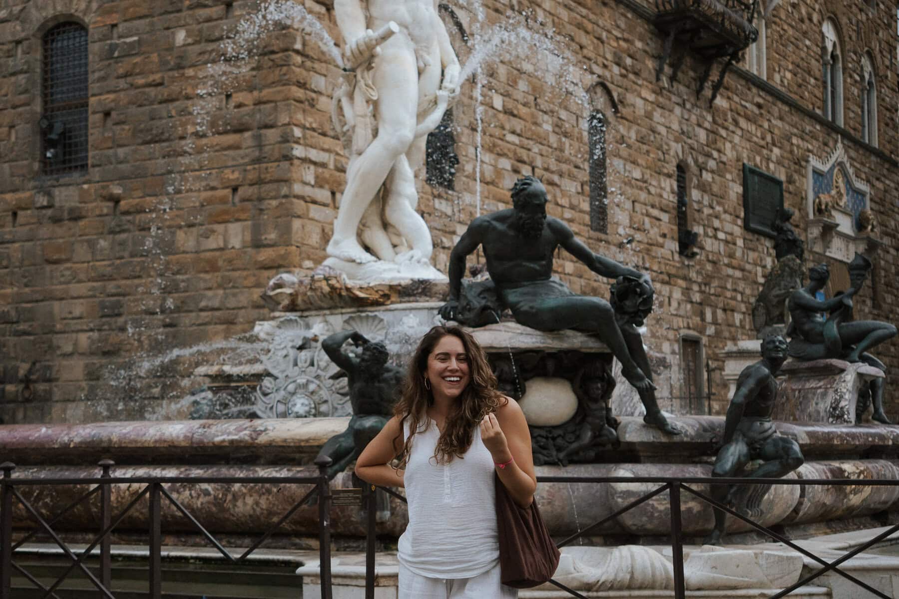 A woman in white stands smiling in front of a historic stone fountain with statues, set against an old brick building—the perfect spot to discover the easiest way to learn Italian and immerse yourself in the local culture.