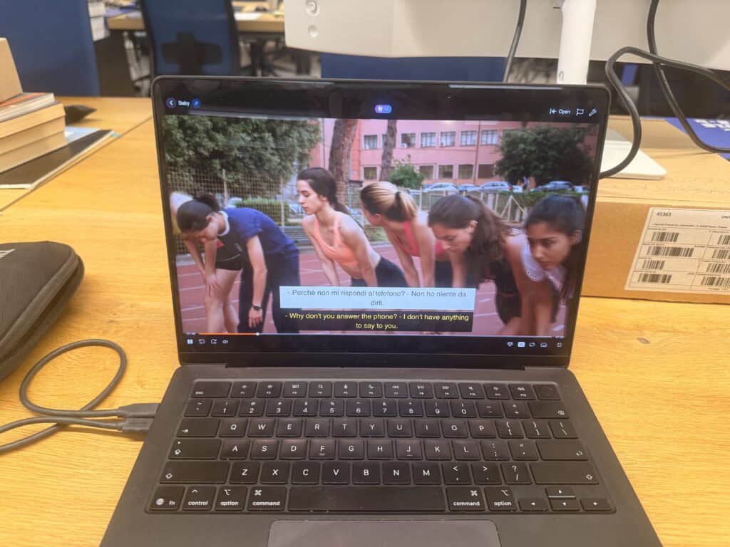 A laptop on a wooden table displays a paused scene from a show with five girls on a track. Italian and English subtitles are visible—the easiest way for an American in Italy to learn Italian while enjoying their favorite series.