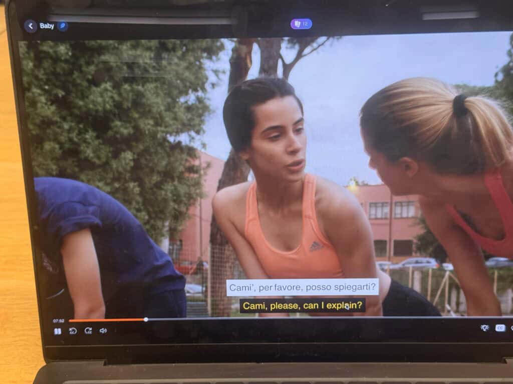 Three young people are outdoors; one woman in an orange tank top is speaking to another woman who is leaning toward her. Subtitles in Italian and English appear at the bottom of the screen, making it the easiest way to learn Italian—especially for any American living in Italy.