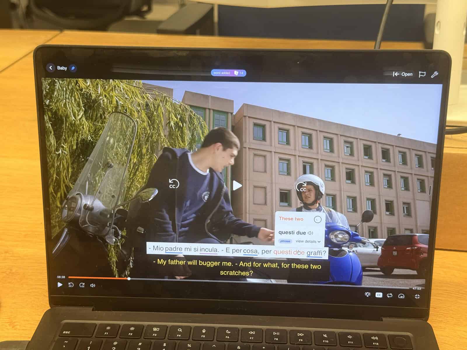 A laptop screen shows a paused video with English and Italian subtitles—an excellent resource for anyone looking to Learn Italian. Two people in jackets are talking near scooters in front of a modern beige building, illustrating the everyday experiences of an American living in Italy and showcasing the easiest way to pick up conversational skills.