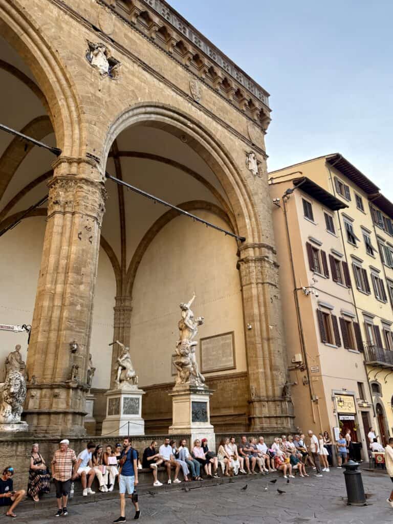 People sit on the steps of the Loggia dei Lanzi in Florence, a highlight for any 3 days in Florence, beneath large stone arches and classical marble statues.