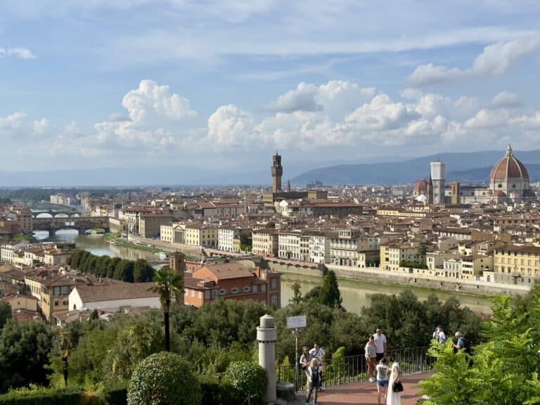 View of the Florence skyline from a high vantage point, showing the Arno River, the Ponte Vecchio, and the Duomo, with people walking on a terrace in the foreground.