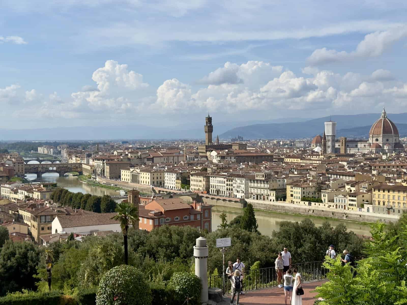 View of the Florence skyline from a high vantage point, showing the Arno River, the Ponte Vecchio, and the Duomo, with people walking on a terrace in the foreground.
