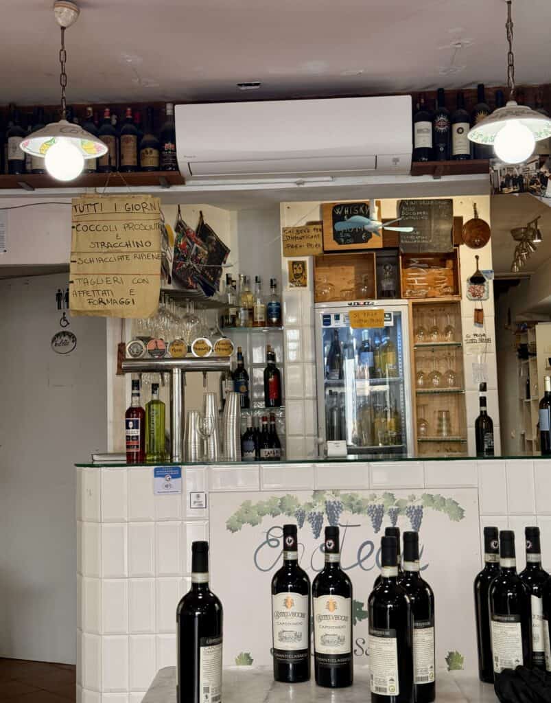 Bar area of a restaurant seen during a 3 days in Florence itinerary, featuring a chalkboard menu, a glass drink cooler, and rows of wine bottles on shelves and the counter.