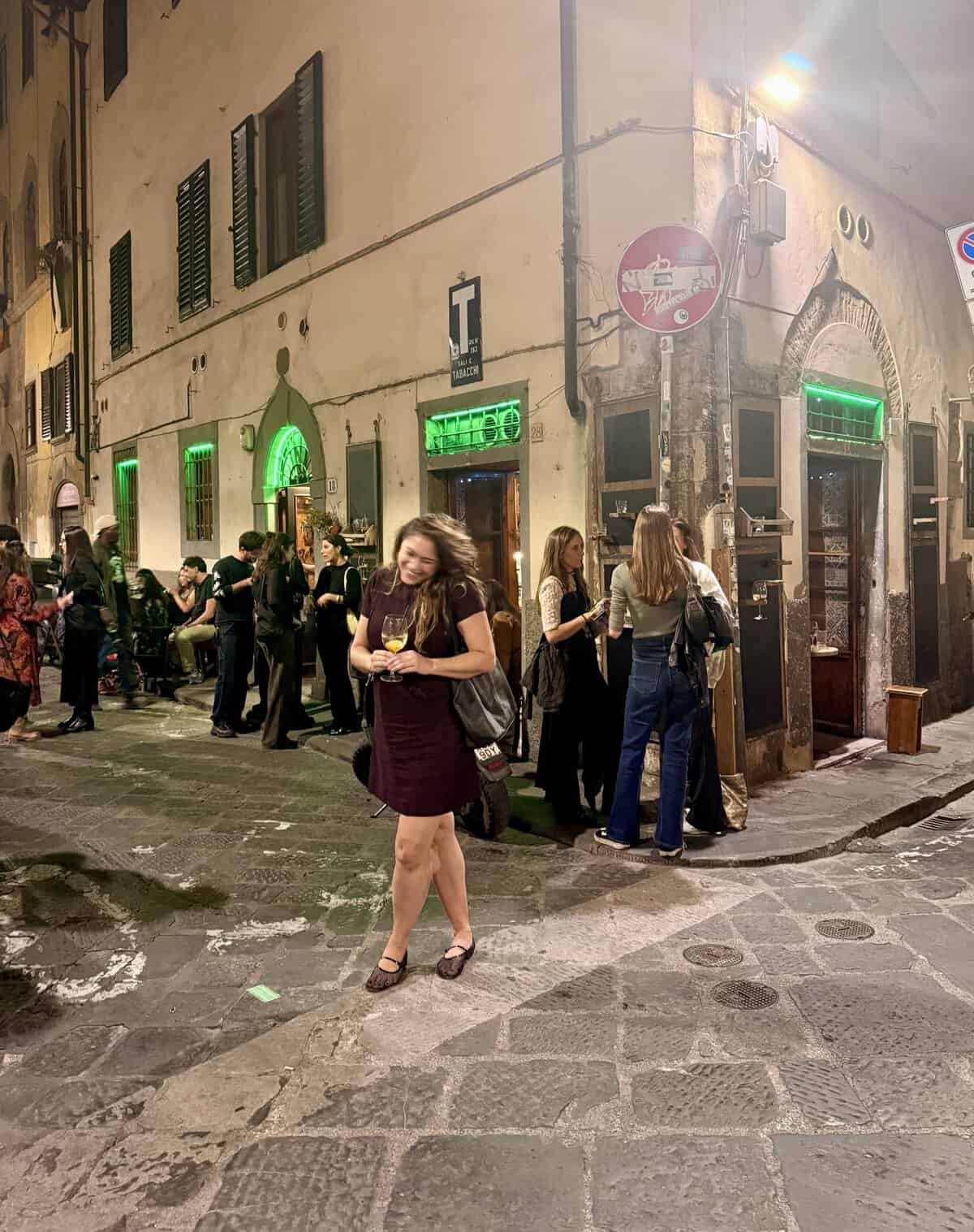 A woman in a dark dress holds a wine glass in a crowded cobblestone street outside a bar at night, a quintessential stop on a 3 days in Florence itinerary.