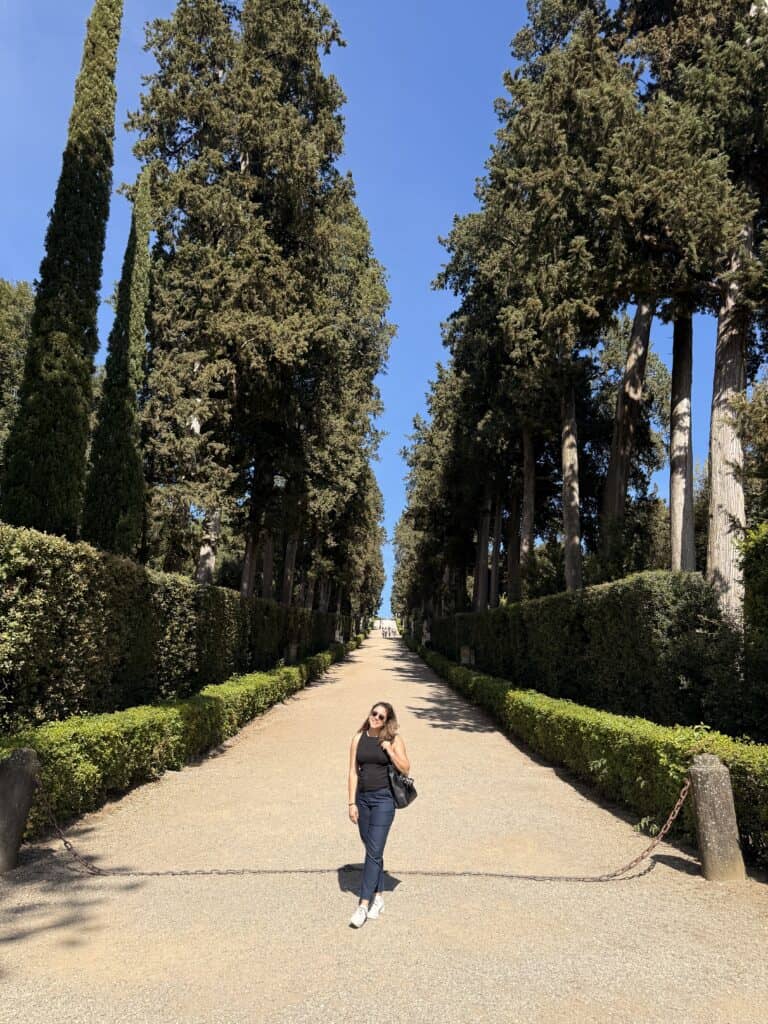 A woman stands on a gravel path lined with cypress trees leading to a building, a serene scene captured during a 3 days in Florence itinerary.