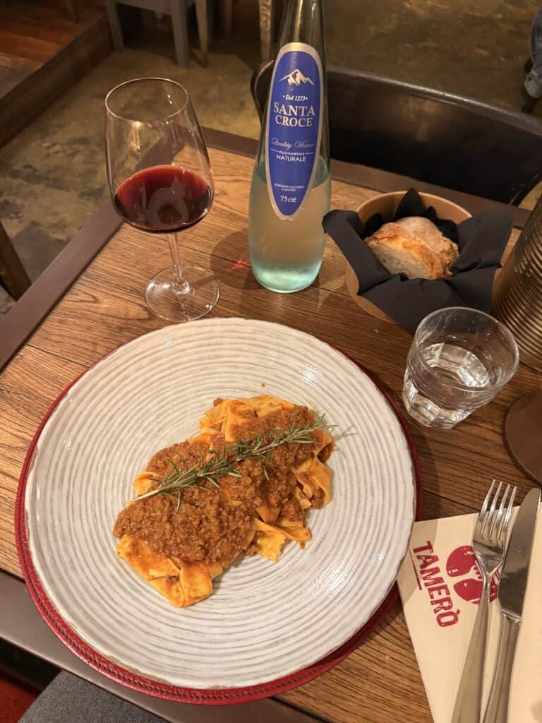 A plate of pappardelle with meat sauce garnished with rosemary, served with wine, bread, and silverware on a wooden table, perfect for your 3 days in Florence.