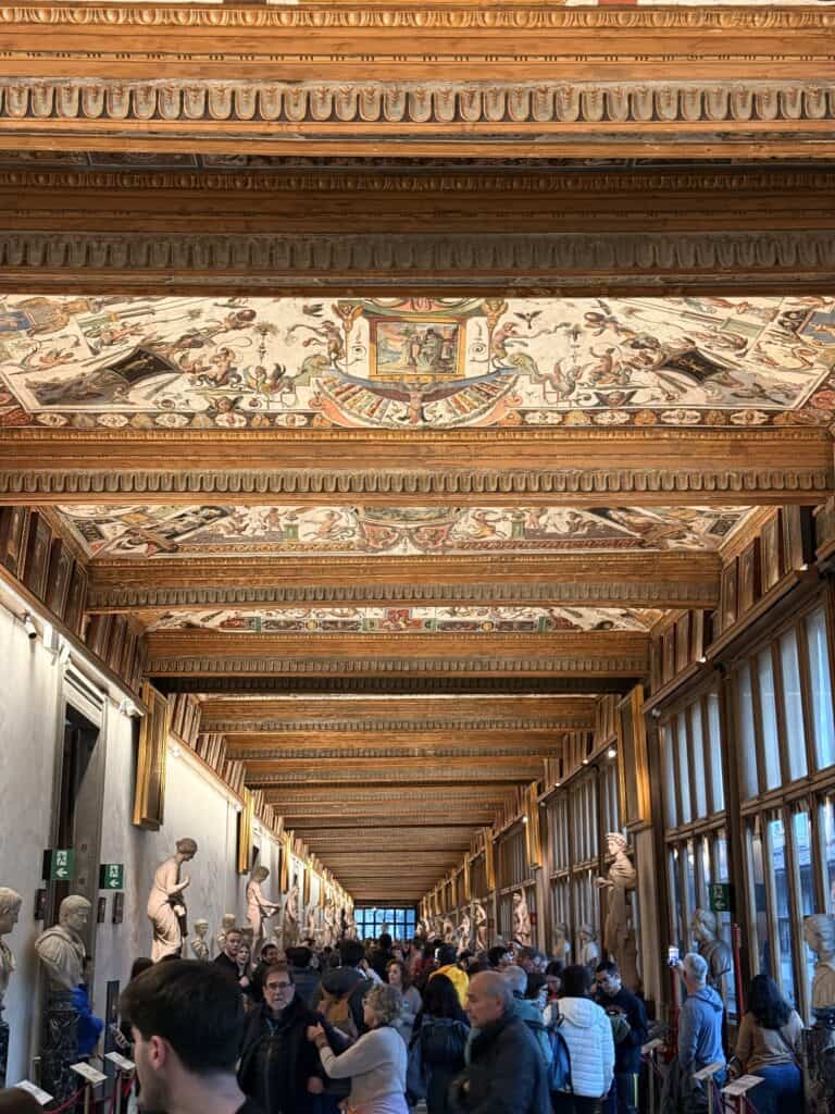 A crowded corridor in the Uffizi Gallery, a highlight of any 3 days in Florence, with vaulted ceilings decorated with frescoes and rows of classical marble statues along the walls.