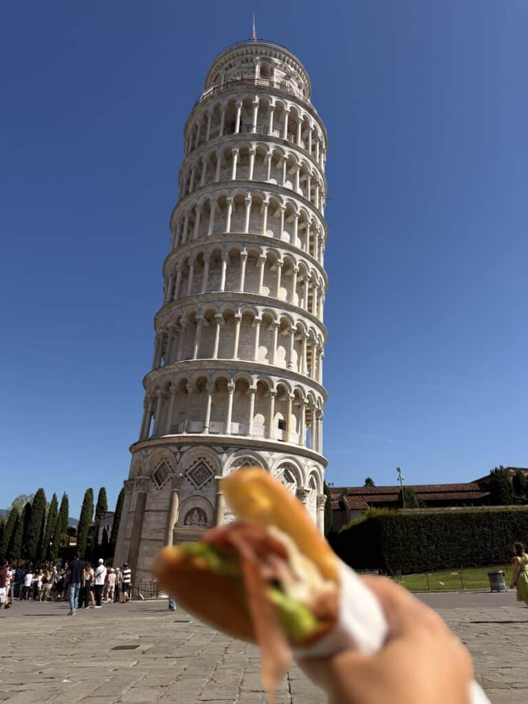 A person holds a sandwich in the foreground with the Leaning Tower of Pisa in the background, a highlight during 3 days in Florence.